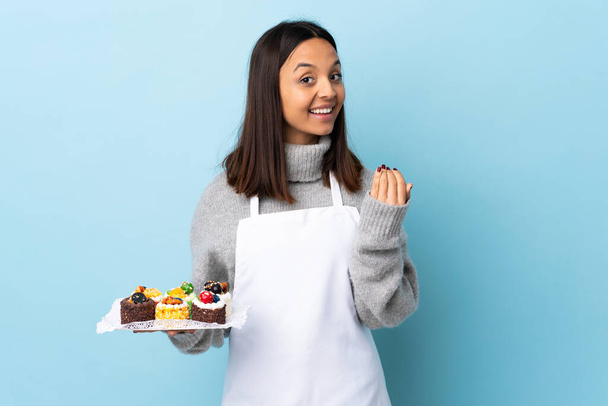Šéfkuchař pečiva drží velký dort přes izolované modré pozadí zvoucí přijít s rukou. Jsem rád, že jsi přišel.. - Fotografie, Obrázek