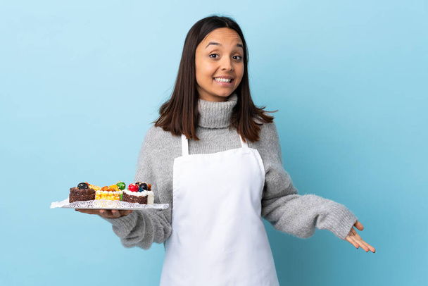 Pastry chef holding a big cake over isolated blue background happy and smiling. - Zdjęcie, obraz