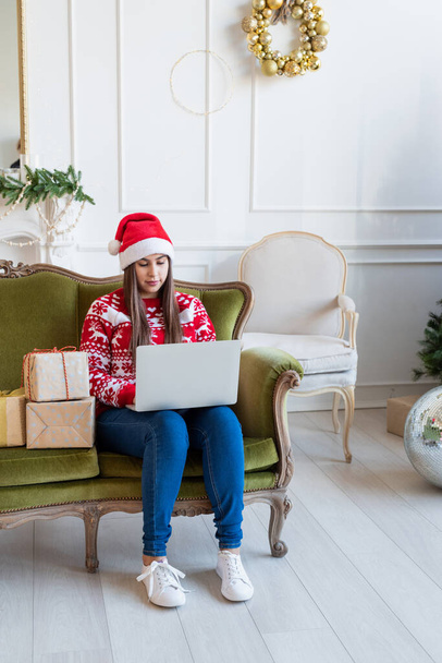 С Рождеством и Новым годом. Молодая брюнетка в шляпе Санты сидит на зеленом диване и работает на ноутбуке в украшенной гостиной - Фото, изображение