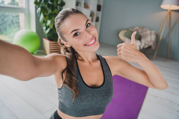 Self-portrait of attractive cheerful sportive confident girl doing physical work out showing thumbup at light home studio indoors - Фото, зображення