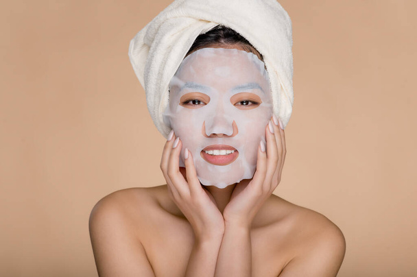 Procédure cosmétique de soins de la peau. Jolie jeune femme asiatique souriante avec un masque hydratant en coton facial sur son visage, et porte une serviette enveloppée de blanc sur la tête, debout sur un fond beige isolé - Photo, image