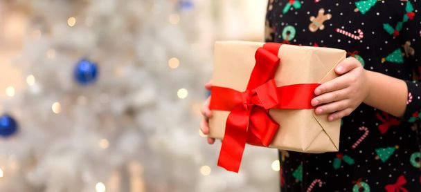 Close-up of a child's hands holding a Christmas gift with a red ribbon.Christmas trees background and bokeh, banner - Фото, зображення