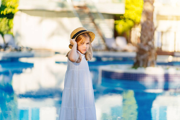 cute little girl in a white dress and straw hat by the pool near the villa.  - Фото, изображение