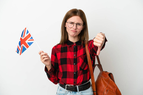 Mavi arka planda izole edilmiş bir Birleşik Krallık bayrağı taşıyan genç bir kadın olumsuz ifadeyle baş parmağını aşağı gösteriyor - Fotoğraf, Görsel
