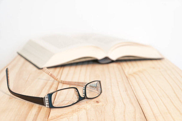Em primeiro plano um par de óculos pretos e brancos, o fundo (fora de foco) um livro em uma mesa de madeira - Foto, Imagem