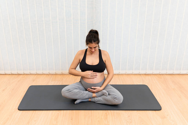 健康な妊娠中の女性座っています上のマット行うヨガタッチ彼女の腹とそれを見て家 - 写真・画像