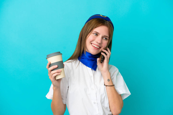 Flugbegleiterin Engländerin isoliert auf blauem Hintergrund mit Kaffee zum Mitnehmen und einem Handy - Foto, Bild