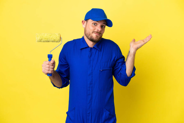 Painter Brazilian man isolated on yellow background having doubts while raising hands - Foto, immagini