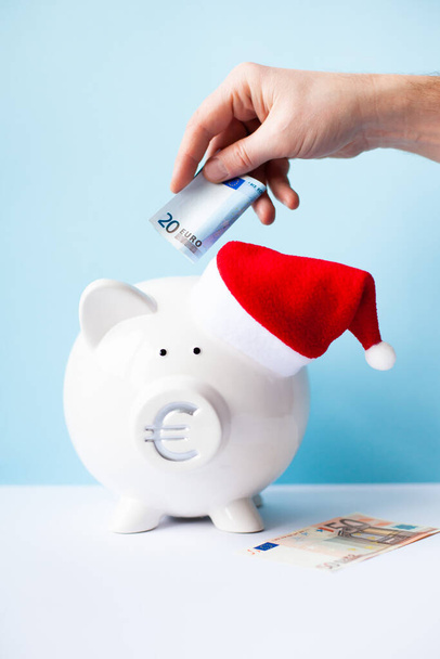 White piggy bank with Santa Claus hat and mans hand puting 20 euro banknote on a blue background. Saving money for Christmas, cash rewards, bonus, sale concept. Vertical image. Selective focus. - Photo, Image