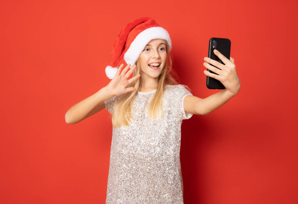 Smiling girl wearing santa hat using smartphone standing isolated over red background. - 写真・画像