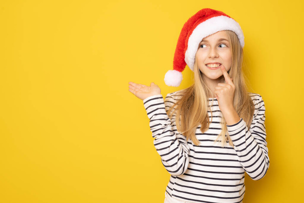 Usmívající se dívka v Santa klobouk ukazující produkt s rukou izolované přes žluté pozadí. - Fotografie, Obrázek