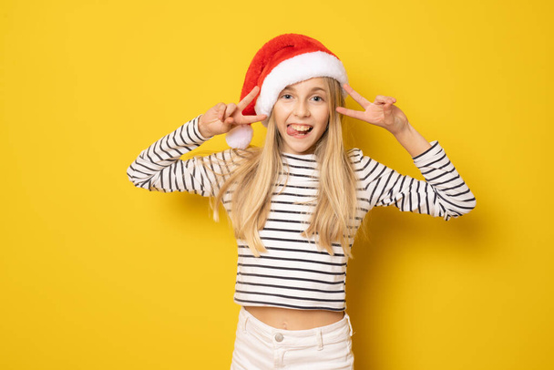 Sarı arka planda izole edilmiş barış sembolü gösteren Noel Baba şapkası takan güzel küçük bir kız.. - Fotoğraf, Görsel