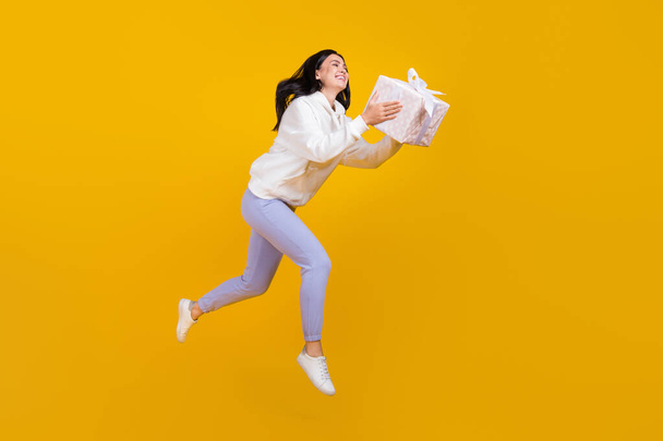 Profile photo of pretty cheerful lady jump hold present box wear white hoodie isolated yellow color background - Fotoğraf, Görsel
