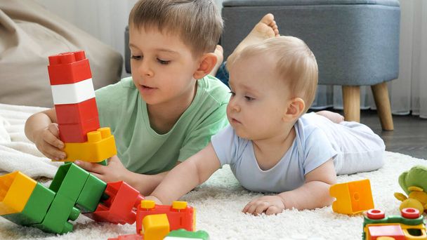 Милый мальчик со старшим братом строит игрушечную башню и играет с игрушками на полу - Фото, изображение