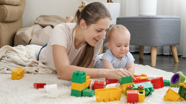 Szczęśliwa uśmiechnięta matka z synkiem budującym zabawkową wieżę z kolorowych cegieł i klocków. Koncepcja rozwoju, edukacji i kreatywności dzieci w domu - Zdjęcie, obraz