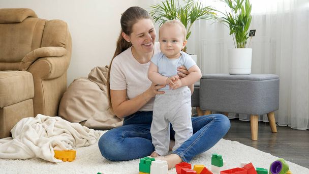 Portrét šťastné usměvavé matky podporující svého malého synka stojící na akrpet v obývacím pokoji - Fotografie, Obrázek