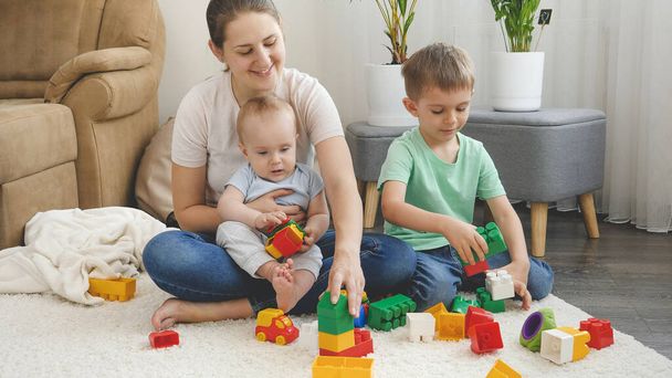 Pikkupoika ja vanhempi veli leikkivät äidin kanssa leluissa matolla olohuoneessa. Käsitys perheen yhteisestä ajasta ja lasten kehityksestä - Valokuva, kuva