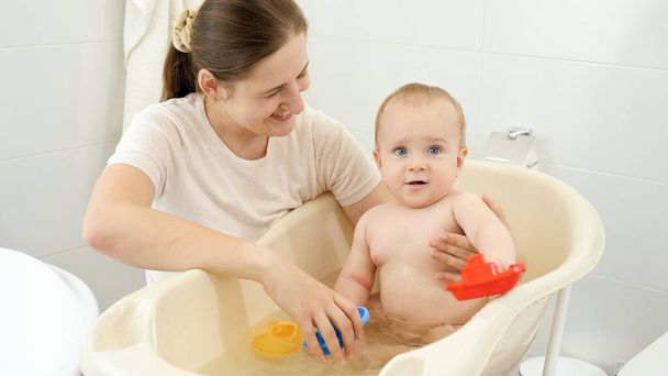 Happy smiling mother and baby boy playing with toy boats and ships in small plastic bath. Concept of children hygiene, healthcare and parenting. - Photo, Image
