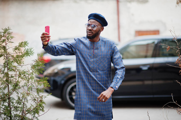 African guy in blue authentic costume, eyeglasses and beret. Fashionable nigerian man near black suv car making selfie on phone. - Photo, Image