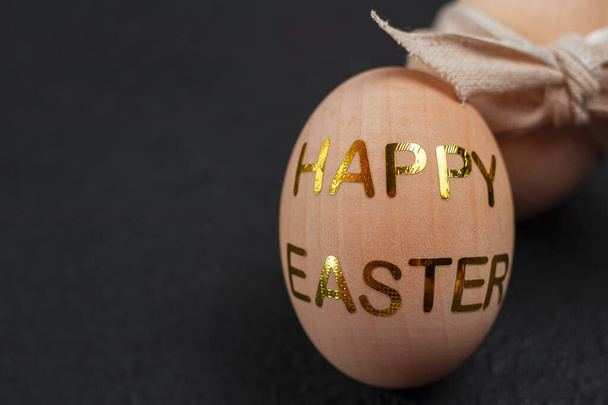 Dřevěná ozdobná vejce se zlatými samolepkami s nápisem HAPPY EASTER. Koncept pohlednice na Velikonoce na černém texturovaném pozadí. - Fotografie, Obrázek