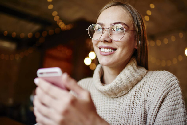 Pleasant woman with toothy smile and eyewear looking away while using smartphone for checking messages on mobile phone in light cafe - Fotoğraf, Görsel
