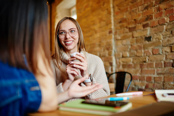 Satisfied female with toothy smile and eyeglasses looking away while sitting at table in cafe with cup of takeaway coffee talking to girlfriend - Photo, Image