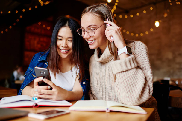 Positive female friends in casual outfits with toothy smiles sitting at wooden table in light cafeteria and browsing mobile phone during break - Foto, imagen