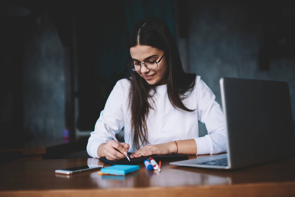 Friendly youthful lady in eyeglasses while sitting at wooden desk in dark workspace with stickers and gadgets while writing down details - 写真・画像