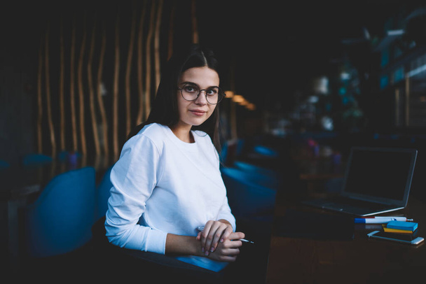 Serious long haired female freelancer in white blouse and spectacles sitting at table with stationery and browsing laptop in cafe - Photo, image