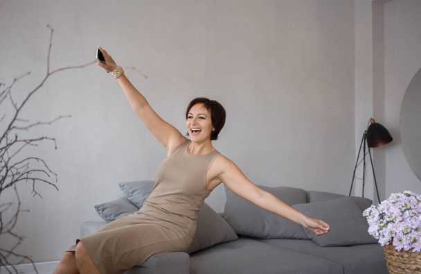 very satisfied beautiful young woman in a tight dress happily spread her arms sits on the couch. joyful elegant business lady celebrating triumph, happy with good news. Positive emotions - Photo, Image