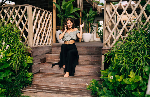 Full body positive barefoot female in stylish clothes emotionally talking on smartphone spreading hand while resting on street sitting on steps of wooden terrace of summer cafe surrounded by plants - Foto, afbeelding