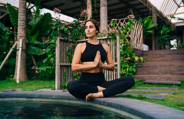 Caucasian woman in black sportswear sitting in lotus pose and looking away while thinking about hatha yoga practice, attractive healthy girl enjoying free time for tranquil enlightenment at patio - Фото, зображення