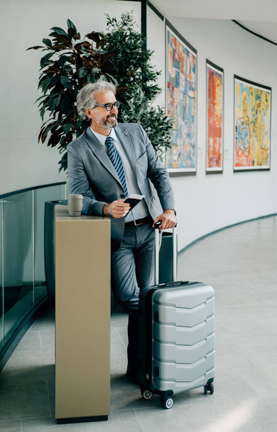 Αναμονή για την πτήση: Ώριμος επιχειρηματίας που κατέχουν διαβατήριο και βαλίτσα, ενώ στέκεται στον τερματικό σταθμό του αεροδρομίου. Χαμογελαστή άνθρωπος ακουμπά σε ένα γραφείο με ένα φλιτζάνι καφέ, ενώ κατέχει διαβατήριο με αεροπορικό εισιτήριο και μεταφορά αποσκευών στο αεροδρόμιο. - Φωτογραφία, εικόνα