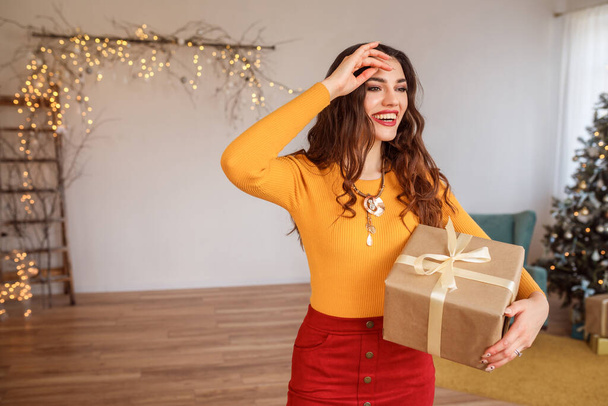 Attractive smiling woman with gift box in her hands celebrating Christmas at home in festively decorated room. - Photo, Image