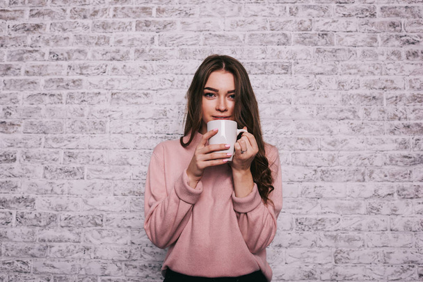 Young calm female with long hair looking at camera standing near brick wall while having rest and drinking tasty coffee - Φωτογραφία, εικόνα