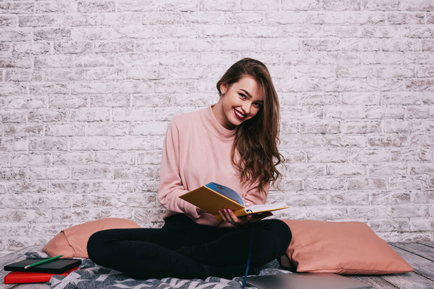 Cheerful female with toothy smile looking at camera sitting with crossed legs with opened book while having free time and reading - Photo, Image