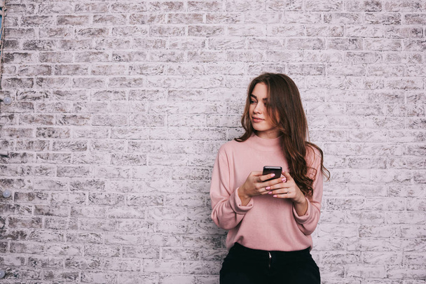 Young smiling female in casual clothes with long hair looking away standing near brick wall and chatting with friends on smartphone - Fotografie, Obrázek