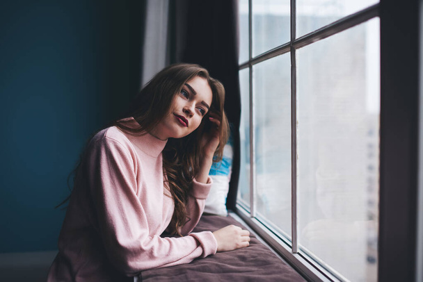 Side view of attractive female in casual clothes with red lips looking away leaning on windowsill and touching hair during rest - Фото, изображение