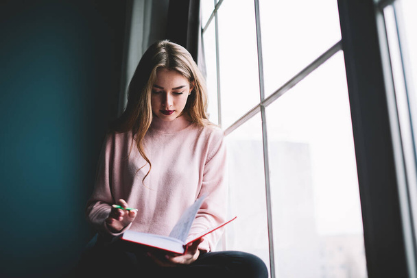 Concentrated female student in casual outfit with long hair sitting near window with pen and notepad and writing notes while preparing for exam - Zdjęcie, obraz