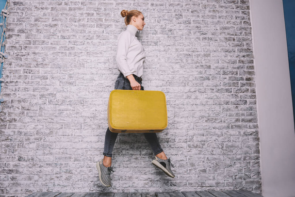 Side view of pensive female model in casual outfit looking away jumping with yellow suitcase on background of brick wall in studio - 写真・画像