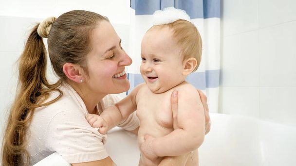 Portrait of smiling baby boy covred in soap foam and mother washing him in bathroom - Photo, Image