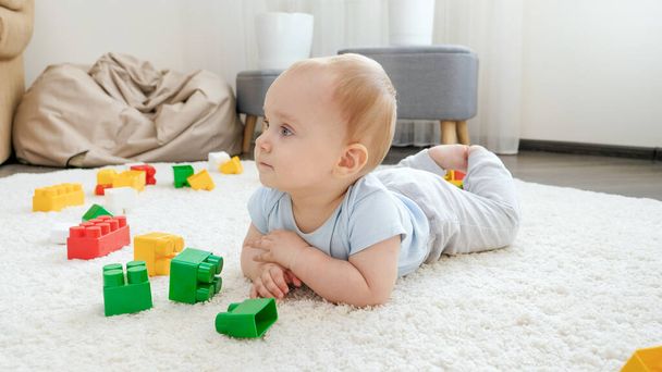 可愛い赤ちゃん男の子はカラフルなおもちゃのレンガやブロックの間でカーペットの上に横たわる - 写真・画像