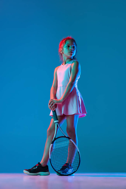 One little girl, beginner tennis player posing isolated on blue studio background in neon light. Sport, study, childhood concept - Foto, afbeelding