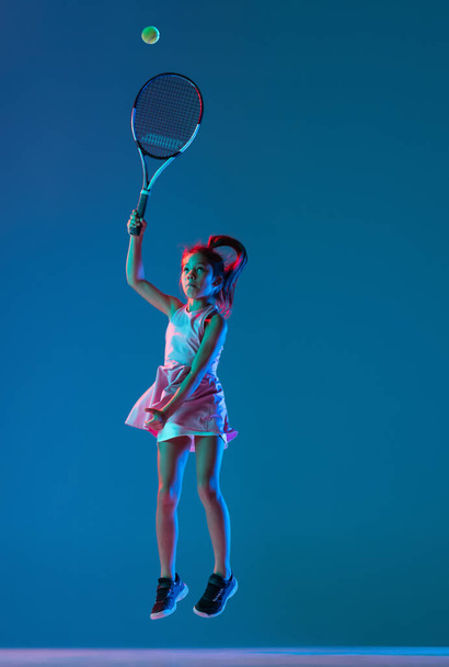 Portrait of little girl, beginner tennis player playing tennis isolated on blue studio background in neon light. Sport, study, childhood concept - Fotografie, Obrázek