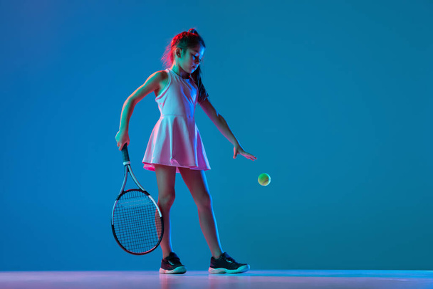 Ein kleines Mädchen, Tennisanfängerin, posiert isoliert auf blauem Studiohintergrund im Neonlicht. Sport, Studium, Kindheitskonzept - Foto, Bild