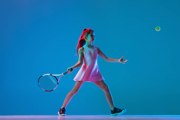 Studio shot of little girl, beginner tennis player training isolated on blue studio background in neon light. Sport, study, childhood concept - Fotó, kép