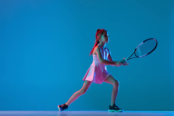 Portrait of little girl, beginner tennis player playing tennis isolated on blue studio background in neon light. Sport, study, childhood concept - Фото, изображение