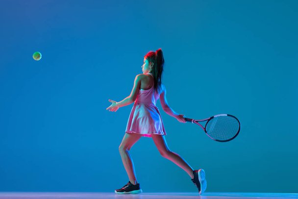 Studio shot of little girl, beginner tennis player training isolated on blue studio background in neon light. Sport, study, childhood concept - Foto, Bild