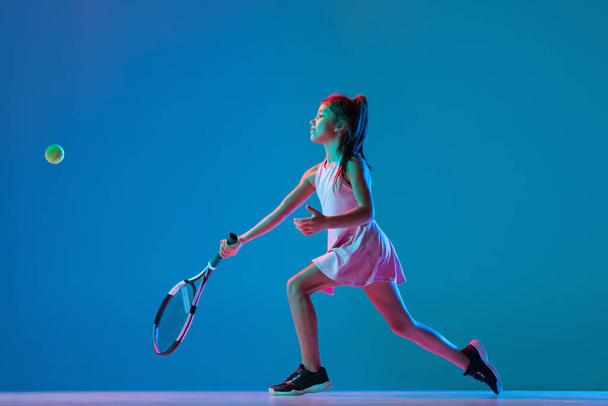 Portrét malé holčičky, začínající tenista hrající tenis izolovaný na modrém pozadí studia v neonovém světle. Sport, studium, koncept dětství - Fotografie, Obrázek
