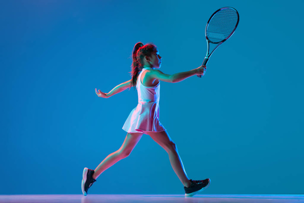 Studio shot of little girl, beginner tennis player training isolated on blue studio background in neon light. Sport, study, childhood concept - Foto, imagen